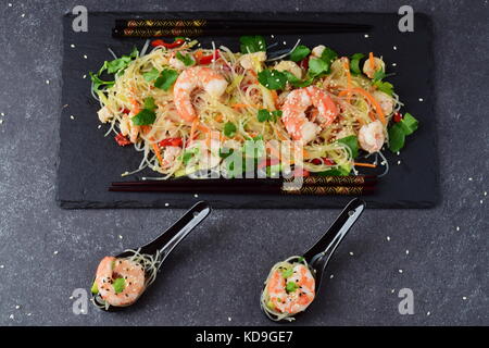 Nudeln mit Garnelen und Gemüse auf einem schwarzen Stein Platte und im traditionellen Suppe löffeln auf einem grauen Abstrakt Hintergrund. gesunde Ernährung Konzept Stockfoto