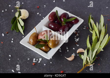 Schwarze und Grüne Oliven in eine weiße Platte auf einem grauen Abstrakt Hintergrund mit Zitrone, Knoblauch, Gewürze. Mediterrane Küche. gesunde Ernährung Konzept Stockfoto