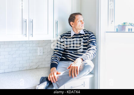 Junge Mann sitzt auf moderne neue Küche Arbeitsplatte durch die Fenster nach außen Denken Stockfoto