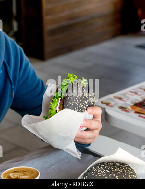 Schwarz Burger mit einem Schnitzel und ein Blatt Salat, Käse, Sesam, in Papier gewickelt, in der Hand eines Mannes Stockfoto