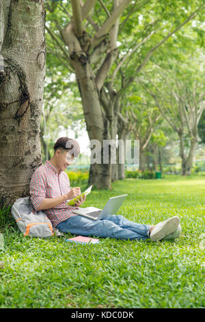 Asiatische student Kompositionen schreiben in Copybooks sitzen auf Park Rasen Stockfoto