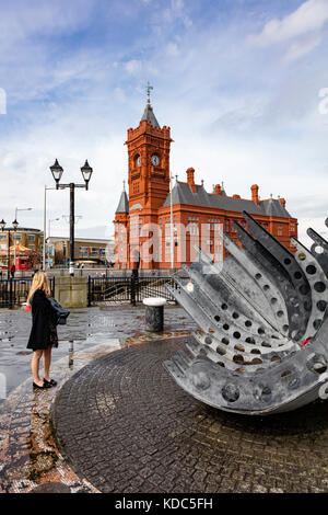 Eine Frau schaut sich das Merchant Seafarer's Memorial und das Pierhead Gebäude an einem kalten, nassen Frühlingstag in Cardiff Bay, Wales, Großbritannien an Stockfoto