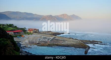 Der Küstenort Hermanus entlang der Garden Route der Sotuh Afrika. Meer Nebel, Wolken Berge schweben. Beruhigende Paradies, jetty Slipanlage Bootshaus Stockfoto