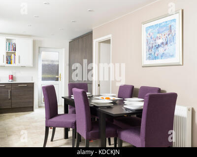 Ein dunkles Holz Esstisch mit violetten Stühlen, für eine Mahlzeit in der modernen Wohnküche eines modernen uk Home eingestellt.