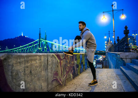 Ein schöner junger Sportler bindet sein Schuhrücken nachts am Flussufer mit einer Brücke hinter sich auf die Straße Stockfoto