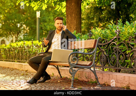 Ein hübscher junger Geschäftsmann Standortwahl auf einer Bank mit seinem Laptop an einem sonnigen Straße neben einem Park, der gerade sein Telefon Stockfoto