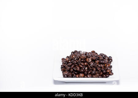Studio Fotografie Bild von gerösteten Kaffeebohnen, close-up-Bild von texturierten braune Bohnen auf einem kleinen Teller auf weißem Hintergrund. Stockfoto