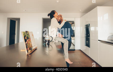 Glückliche junge Frau sitzt auf der Arbeitsplatte in der Küche und umarmt ihren Mann stehen. Liebevolle junge Paar am Morgen in der Küche. Stockfoto