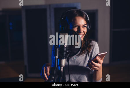 Lächelnd weibliche Wiedergabe Sänger mit Mikrofon und Handy im Aufnahmestudio. Frau mit der Aufnahme eines Songs für Ihr Album im Music Studio. Stockfoto