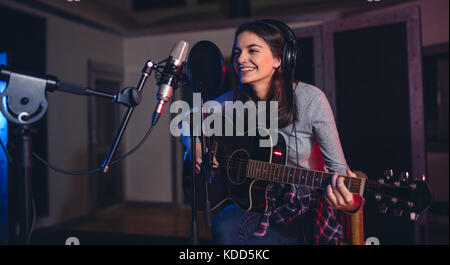 Glückliche junge Frau, die mit der Aufnahme eines Songs in einem professionellen Studio. Lächelnd weibliche sitzen auf der Vorderseite des Mikrofons mit Gitarre im Studio. Stockfoto