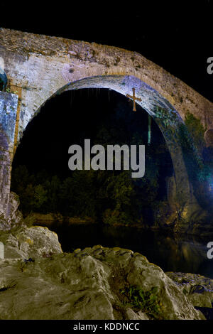 Hängende Kreuz unter der alten römischen Brücke (Puente Romano), 13.jahrhundert, über den Fluss Sella in der Nacht in Cangas de Onis. Asturien. Spanien. Stockfoto