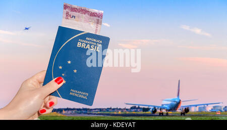 Hand mit einem brasilianischen Reisepass mit 5 Reais Hinweis im Inneren, mit Flugzeugen im Fokus Hintergrund bei Sonnenuntergang Stockfoto