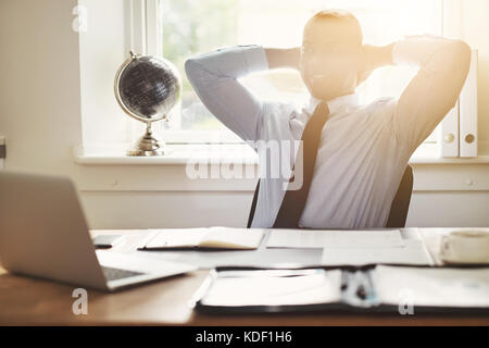 Lächelnden jungen afrikanischen Geschäftsmann in einem T-Shirt und souverän lehnte sich in seinem Stuhl zurück in seine Hände hinter den Kopf in seinem Büro Schreibtisch binden Stockfoto