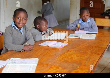 Drei afrikanische Schuljungen in Uniform sitzen an einem Schreibtisch und studieren, ein Junge schaut lächelnd in die Kamera Stockfoto