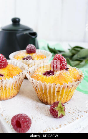 Frisch gebackene Muffins mit Himbeeren, dekoriert mit Puderzucker, Teekanne auf einem weißen Holz- Hintergrund. Stockfoto