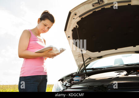 Junge Frau mit Buch vorne am Auto mit offener Haube Stockfoto