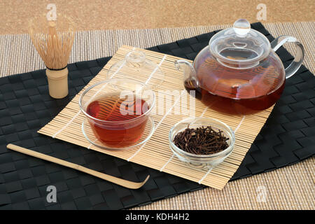 Gesunde japanische kuchika geröstete Zweig Tee mit Glas Teekanne und Tassen, Rührer und Schneebesen auf Bambus Hintergrund. hat viele gesundheitliche Vorteile. Stockfoto