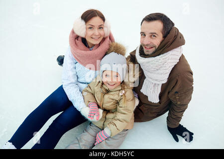 Junge Eltern und ihre Tochter in winterwear auf Kamera beim Sitzen auf der Eislaufbahn Stockfoto