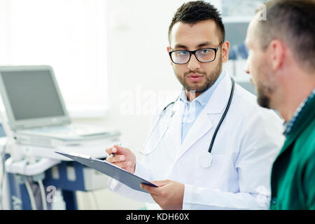 Yougn Doktor in weiß und Brille erklärt seinem Patienten, wie man neue Medikamente nimmt Stockfoto