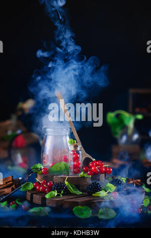 Jar mit frischen Beeren, Johannisbeere, und Blackberry mit Holzlöffel und steigenden Rauch auf einem schwarzen Hintergrund. Labor und Wissenschaft Konzept. magische Stil Stockfoto