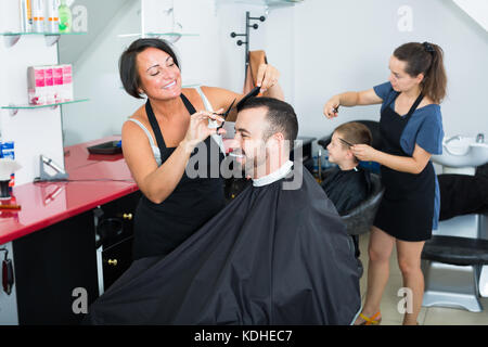 Positive Frau Friseur mit Instrument schneiden Lächeln männlichen Klienten in überfüllten Beauty Salon Stockfoto