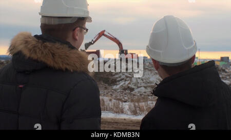 Rückansicht von Architekten, die Baustelle inspizieren. Zwei Ingenieure in Schutzhelmen auf Baustellenbaggern im Hintergrund. Ingenieure prüfen Dokumente auf der Baustelle gegen Bagger. Stockfoto