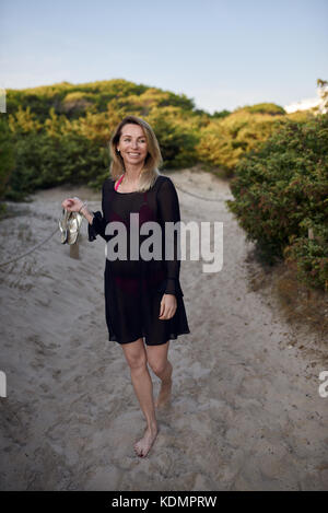 Attraktive schlanke blonde Frau ihre Schuhe tragen als sie Spaziergänge an einem Sandstrand weg Lächeln glücklich an der Seite im Sommerurlaub Stockfoto