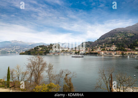 Laveno, Lago Maggiore, Italien. Malerische Aussicht auf die Promenade, die zum Hafen führt, die Fähre und der Kirche. Im Hintergrund der Umgebungslichtsensor Stockfoto