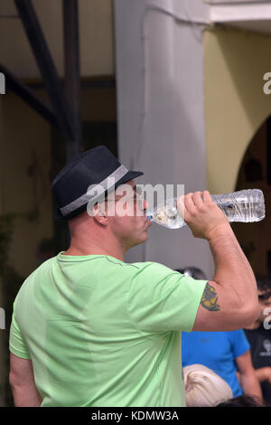 Ein Mann das Trinken aus einer Flasche Wasser mit einem Trilby Hut und Schwitzen ausgiebig nach einem Rucksack mit einem Schweiß befleckt Shirt nass und Patches Stockfoto