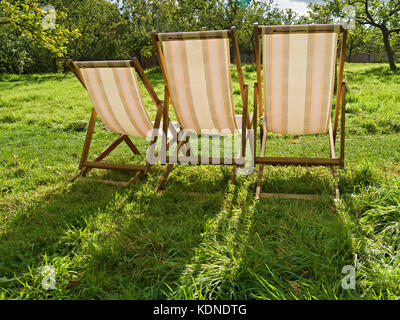 Drei Liegestühle aus Holz und Leinwand mit Hintergrundbeleuchtung und langen Schatten auf grünem, grasbewachsenem Garten, Sommer, Großbritannien Stockfoto