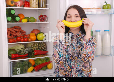 Ein junges Mädchen hält eine Banane in den Mund, ein Lächeln nachahmen. schönen jungen Mädchen in der Nähe der Kühlschrank. Stockfoto