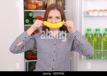 Ein junges Mädchen hält eine Banane in den Mund, ein Lächeln nachahmen. schönen jungen Mädchen in der Nähe der Kühlschrank. Stockfoto