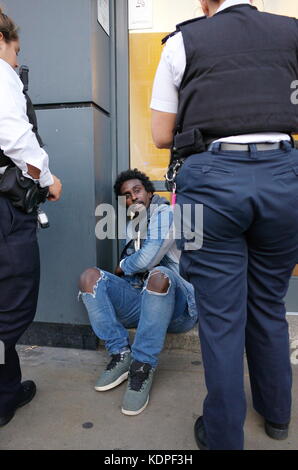 Die somalische Mann in Handschellen sitzt auf dem Boden, während zwei weibliche Polizisten über ihm stehen. London, England, Großbritannien Stockfoto