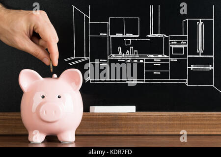 Nahaufnahme der Person einfügen Münze in Sparschwein mit Küche Bild auf der Tafel im Hintergrund Stockfoto