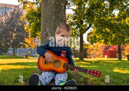 Kleiner Junge Holding akustische Gitarre während der Sitzung lehnte sich an einen Baum Stockfoto