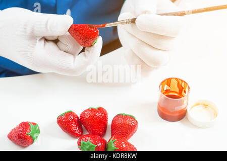 Nahaufnahme einer weiblichen Konditor in blauen Uniform und medizinische Handschuhe malt eine Holz- Pinsel mit roter Farbe eine Erdbeere Schokolade, auf einem weißen Tisch liegen Stockfoto