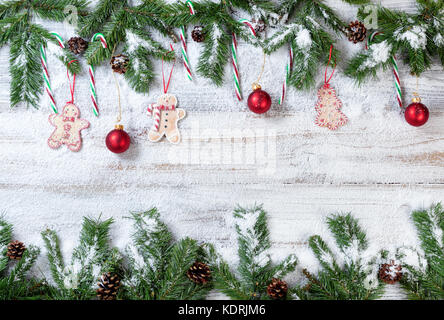 Snowy christmas immergrüne Zweige mit hängenden roten Ornamenten, Cookies und Zuckerstangen auf rustikalen weiß Holz Hintergrund Stockfoto