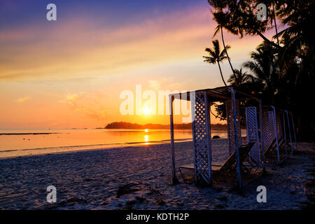 Schönen goldenen Sonnenaufgang über den Sandstrand auf der Insel Bintan in Indonesien mit Silhouette von Liegestühlen und Kokospalmen mit kopieren. Stockfoto