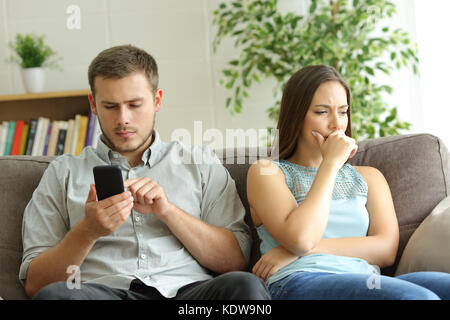 Ehemann zu smart phone Sie Inhalt neben seinem besorgt, Frau, sitzen auf einem Sofa zu Hause süchtig Stockfoto