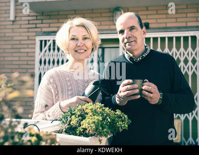 Senior Paar auf einer Terrasse in der Nähe ihrer Heimat. reife Frau hält im Garten Zubehör und Mann ist Tee trinken aus einem Becher Stockfoto