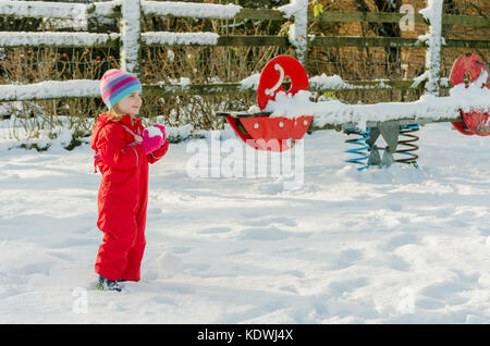 Cute Vorschüler kleines Mädchen im Winter schnee Szene das Tragen der roten Schneeanzug Holding einen Schneeball. Aktiven Lebensstil im Freien. Stockfoto