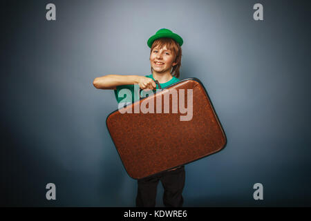 Europäische Erscheinungsbild Teenager Junge in ein Shirt in grünen Hut hält einen alten Koffer in der Hand auf grauem Hintergrund, Abenteuer cross Prozess Stockfoto