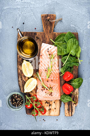 Zutaten für gesundes Essen zu kochen. Rohes Lachsfilet, Spinat, Tomaten, Olivenöl, Zitronensaft, Pfeffer, Rosmarin und Gewürze auf einem rustikalen Holzbrett Stockfoto