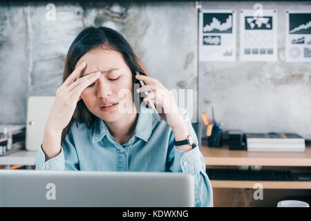Betonte asiatischen kreative Designer Frau, die ihr Gesicht mit der Hand und fühlen sich verärgert während Gespräch auf dem Handy mit Kunden vor Laptop auf Stockfoto