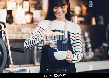 Asien Frau barista gießen Sie Milch in die heiße Tasse Kaffee an der Bar vor der Maschine im Cafe Restaurant, Essen Geschäftsinhaber Konzept Stockfoto