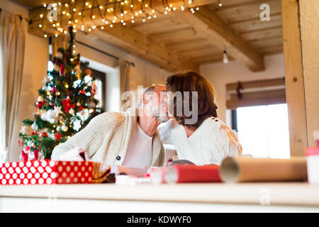 Schönes Paar in weißen wollenen Pullover am Tisch sitzen Verpackung Weihnachtsgeschenke zusammen. senior Paar küssen. Stockfoto