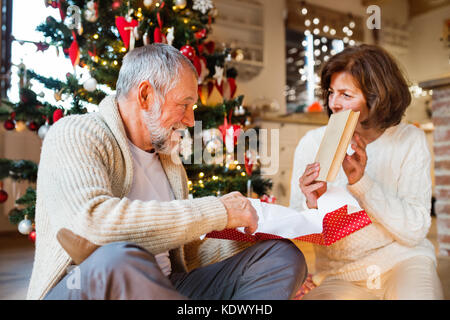 Senior Paar sitzt auf dem Boden vor der beleuchteten Weihnachtsbaum in Ihrem Haus schenken, Frau ihr Geschenk auspacken. Stockfoto