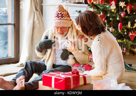 Älterer Mann sitzt auf dem Boden vor der beleuchteten Weihnachtsbaum mit seiner Frau. der Mann, der versucht, auf gestrickte Schal, Mütze und Handschuhe. Stockfoto