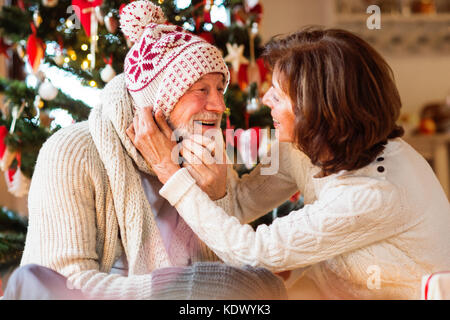 Älterer Mann sitzt auf dem Boden vor der beleuchteten Weihnachtsbaum mit seiner Frau. der Mann, der versucht, auf gestrickte Schal, Mütze und Handschuhe. Stockfoto