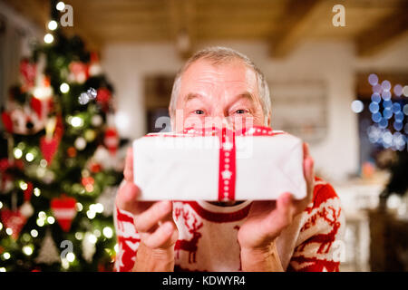 Ältere Mann, der vor der beleuchteten Weihnachtsbaum in seinem Haus Holding, Gefühl zufrieden. Stockfoto